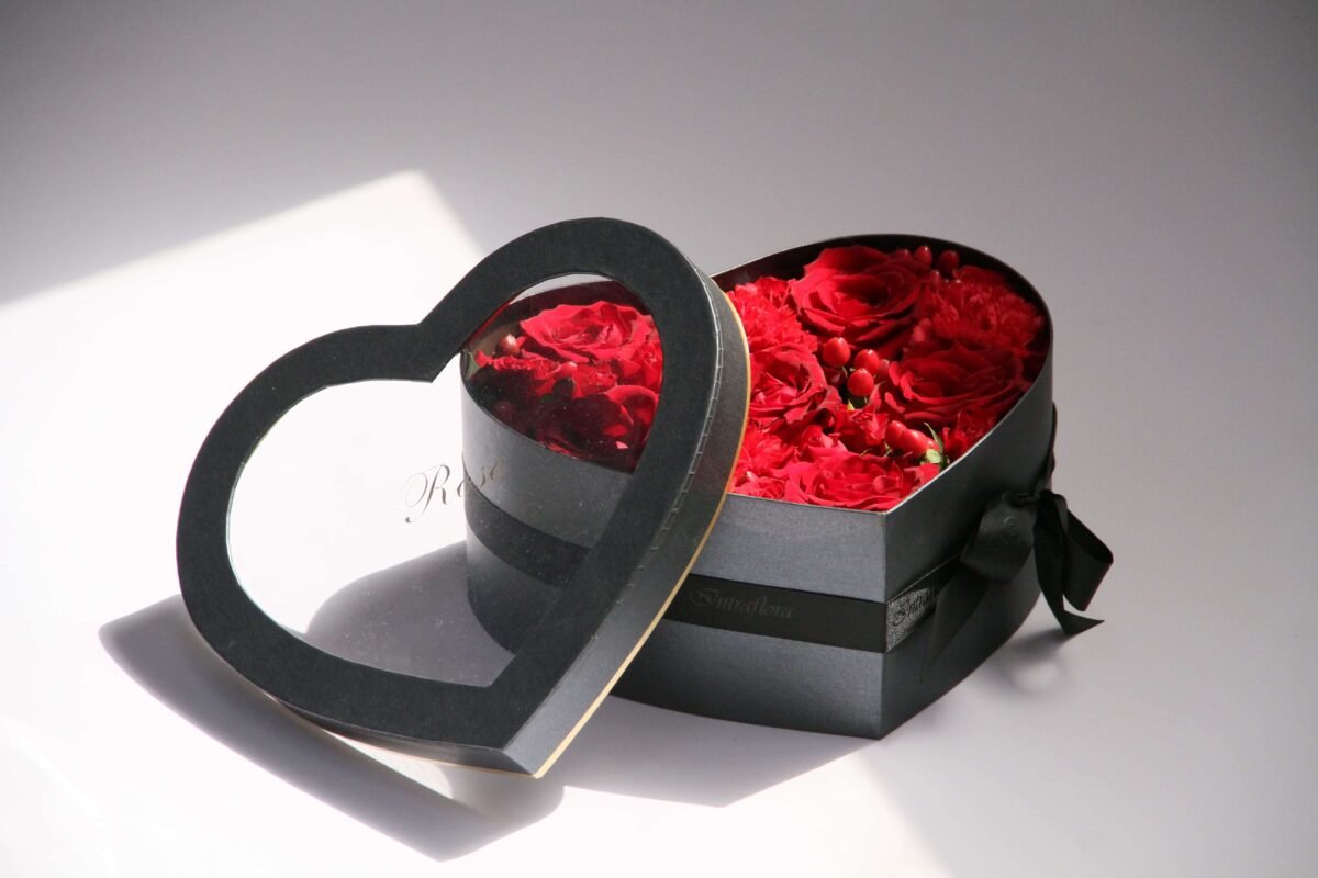 A dark gray, heart-shaped box with a transparent lid, showcasing red roses inside. The box also features a black ribbon and a small tag with the word "Rose" on it.
