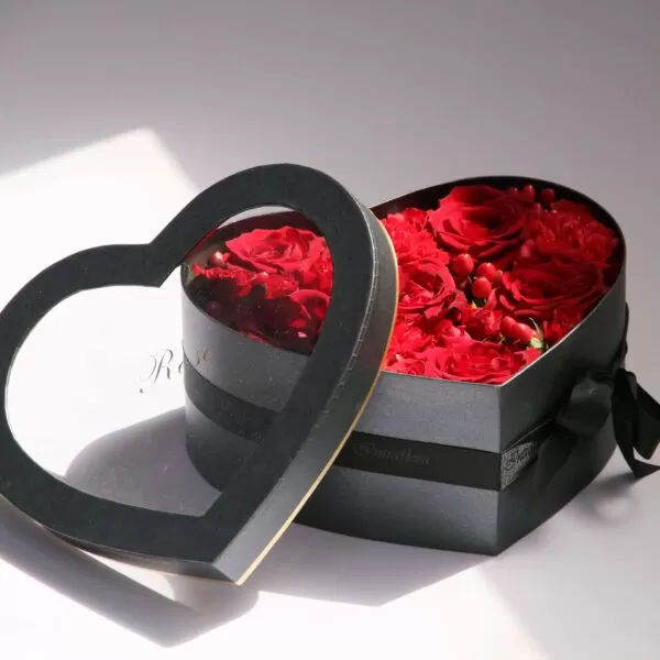 A dark gray, heart-shaped box with a transparent lid, showcasing red roses inside. The box also features a black ribbon and a small tag with the word "Rose" on it.
