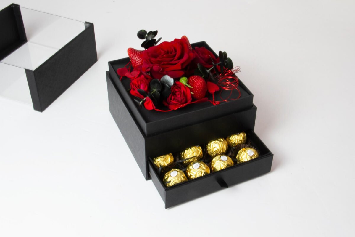 A black square box with a smaller black square box stacked on top. The top box contains red roses, red strawberries, and black decorative twigs. The bottom box has a pull-out drawer containing gold-wrapped chocolates.