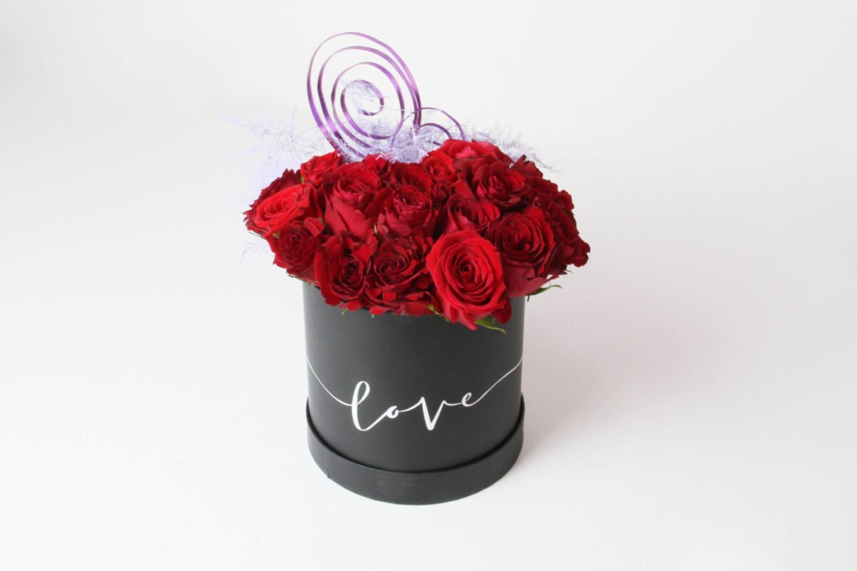 A black, cylindrical flower box filled with dark red roses, with the word "love" written in white script on the side. The box also features purple decorative twigs and light purple feathery decorations, against a light background.