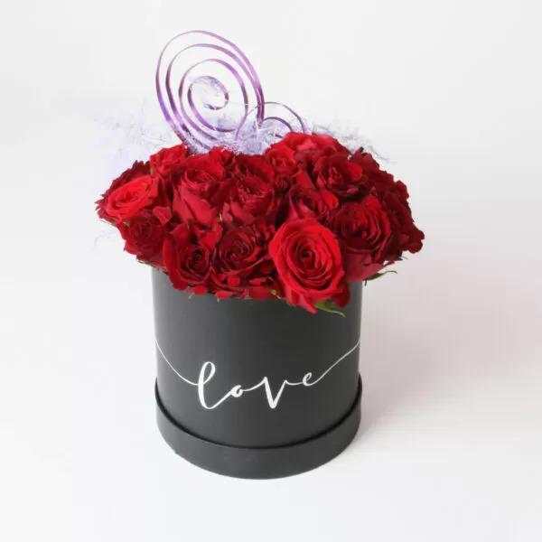 A black, cylindrical flower box filled with dark red roses, with the word "love" written in white script on the side. The box also features purple decorative twigs and light purple feathery decorations, against a light background.