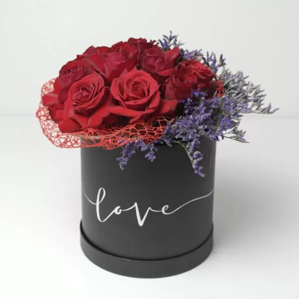 A black, cylindrical flower box filled with dark red roses and purple waxflowers, with the word "love" written in white script on the side. The box also features a red mesh decoration, against a light background.