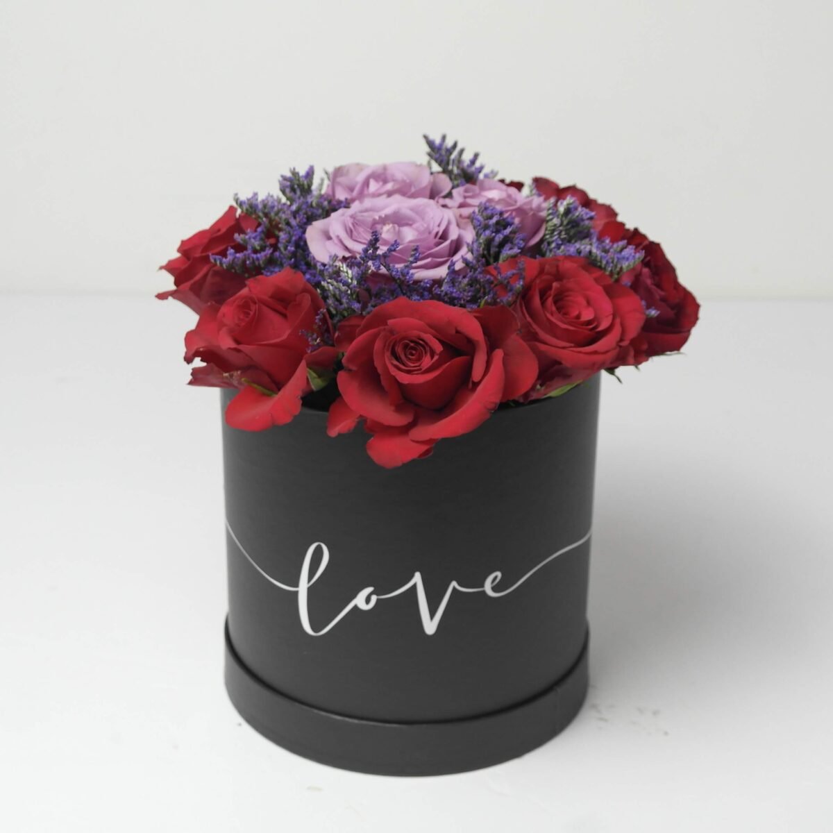 A black, cylindrical flower box filled with dark red roses, purple roses, and purple waxflowers, with the word "love" written in white script on the side, against a light background.