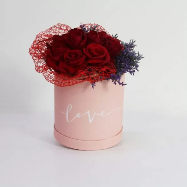 A pink, cylindrical flower box filled with dark red roses and purple waxflowers, with the word "love" written in white script on the side. The box also features a red mesh decoration, against a light background.