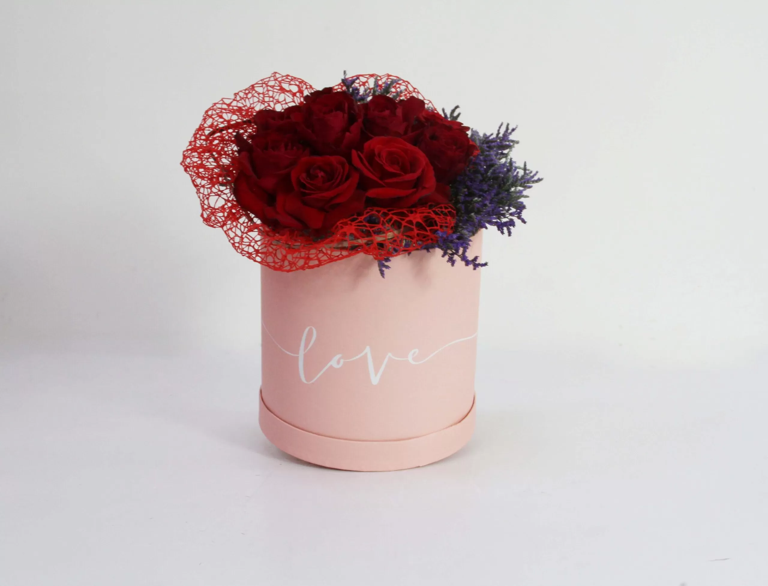 A pink, cylindrical flower box filled with dark red roses and purple waxflowers, with the word "love" written in white script on the side. The box also features a red mesh decoration, against a light background.