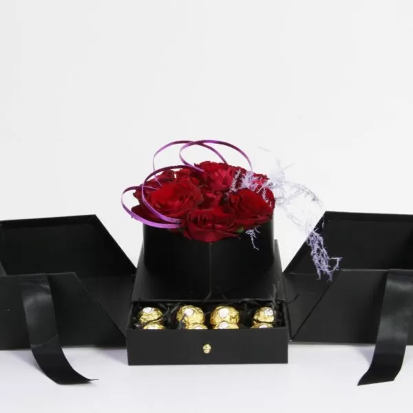 A black square box with a smaller black square box inside, filled with dark red roses and purple decorative twigs. The larger box has four flaps that open to reveal a small drawer containing four gold-wrapped chocolates. The box also features a small tag with purple decorations.