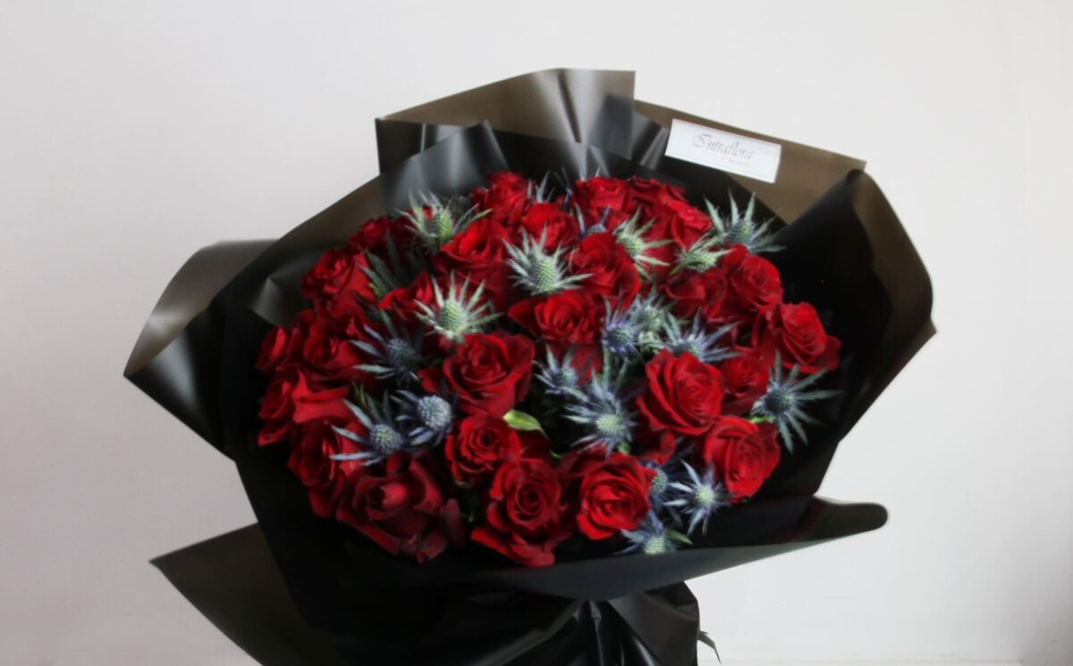A bouquet of red roses and blue thistle flowers, wrapped in black paper with a white label, against a light background.