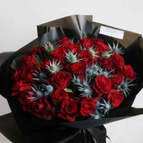 A bouquet of red roses and blue thistle flowers, wrapped in black paper with a white label, against a light background.