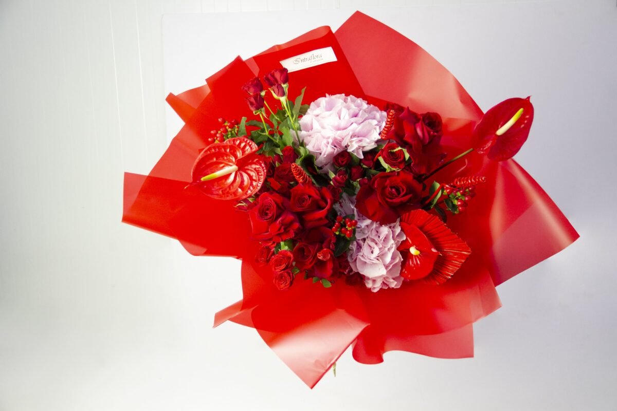 A bouquet of red roses, red anthuriums, pink hydrangeas, red tulips, and red berries, wrapped in bright red paper, against a light background. The bouquet also features a small white label with the word "Smiles" on it.