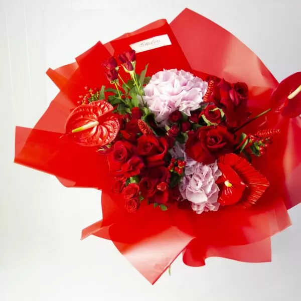 A bouquet of red roses, red anthuriums, pink hydrangeas, red tulips, and red berries, wrapped in bright red paper, against a light background. The bouquet also features a small white label with the word "Smiles" on it.