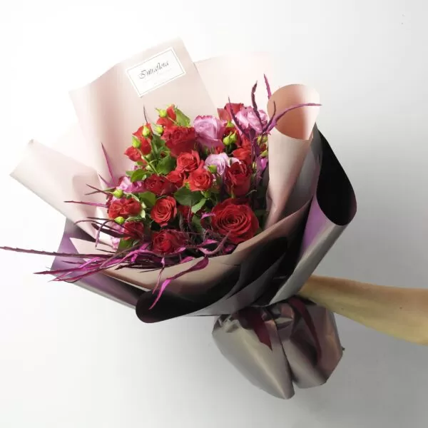 A hand holding a bouquet of red roses and two-tone eustoma flowers, wrapped in pink and purple paper, against a light background. The bouquet also features purple twigs and a small "Intraflora" logo sticker.