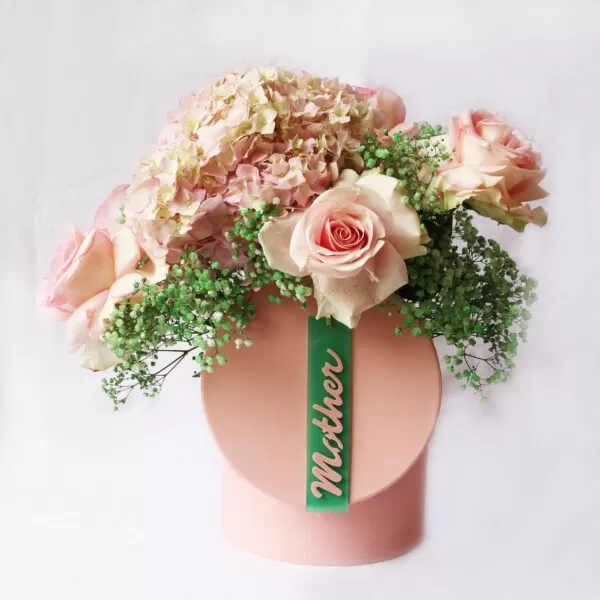 A pink cylindrical box with a rounded top, containing pink hydrangeas, light pink garden roses, and white gypsophila flowers, decorated with a green ribbon that says "Mother" in cursive script, against a white background.