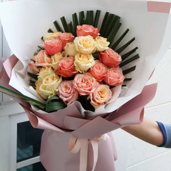 A close-up shot of a bouquet featuring pink and yellow roses, accented with green leaves, wrapped in white and pink paper and tied with a light pink ribbon, being held by a person's arm wearing a blue shirt.