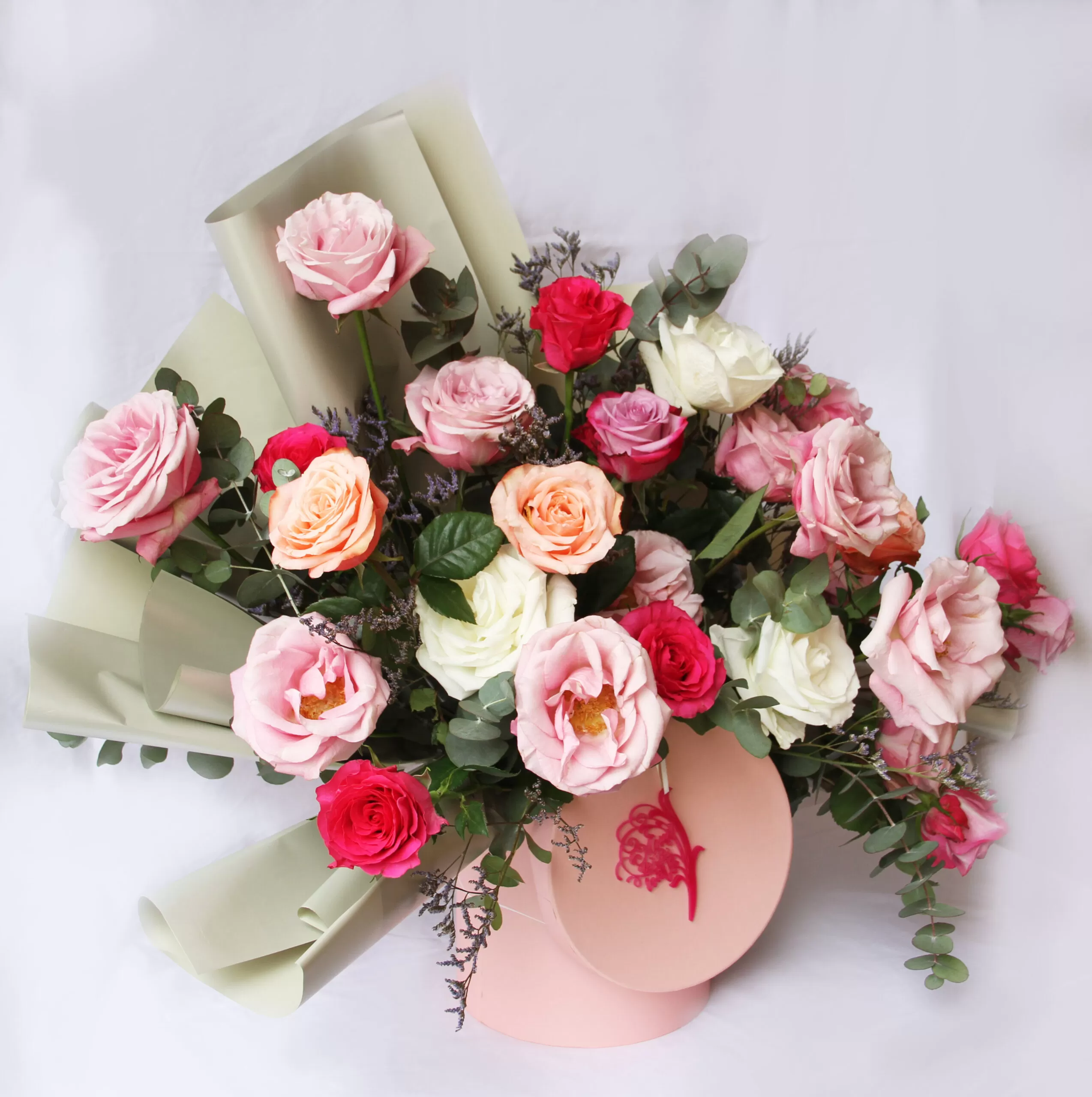 Overhead shot of a bouquet featuring a variety of roses in shades of pink, peach, red, and white, arranged with greenery and wrapped in a light green paper, tied with a pink ribbon featuring a small illustration.