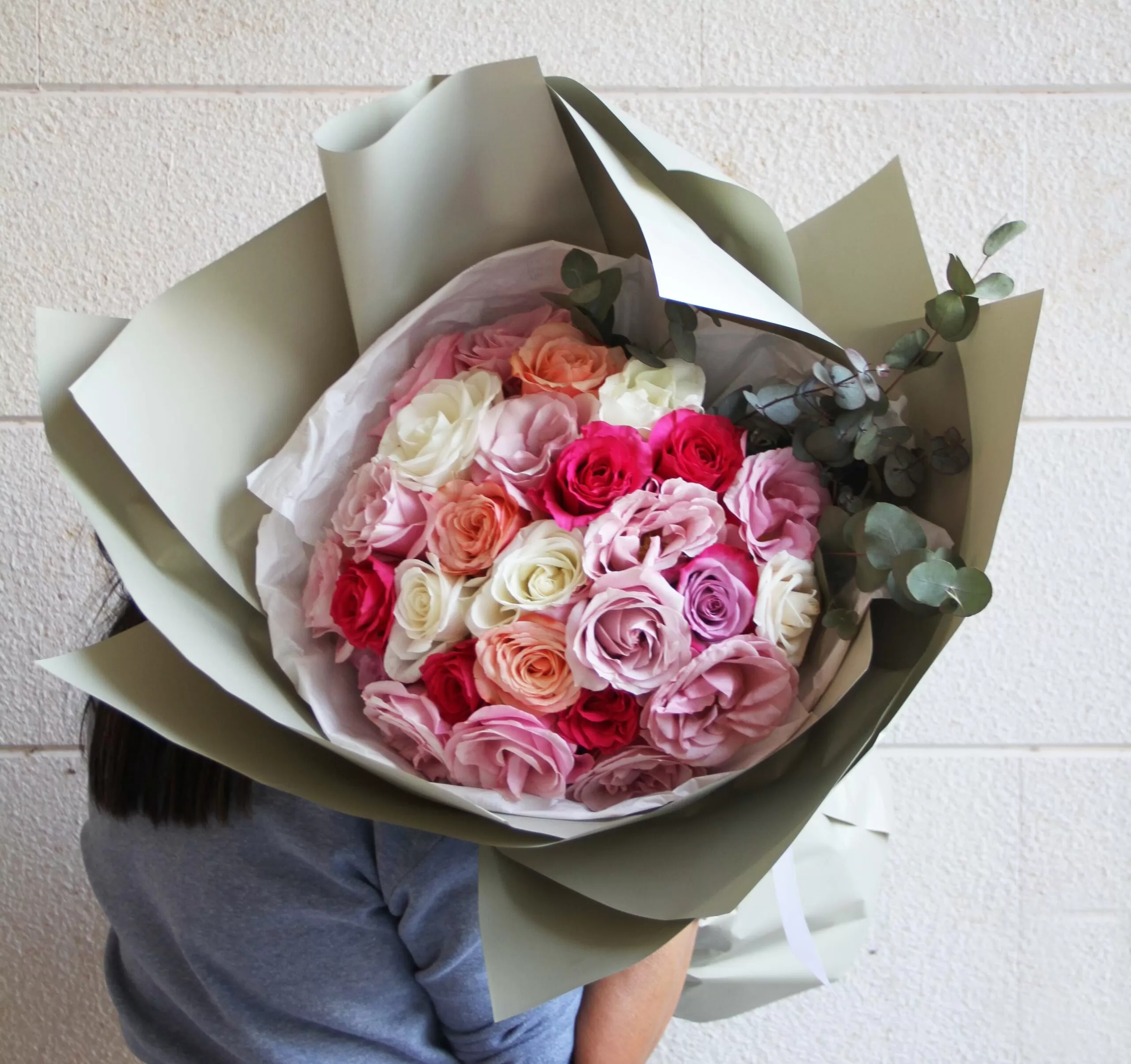 A close-up shot of a person wearing a gray shirt holding a large, round bouquet of roses in various colors including pink, peach, red, white, and purple, wrapped in light green paper with greenery visible on the side.