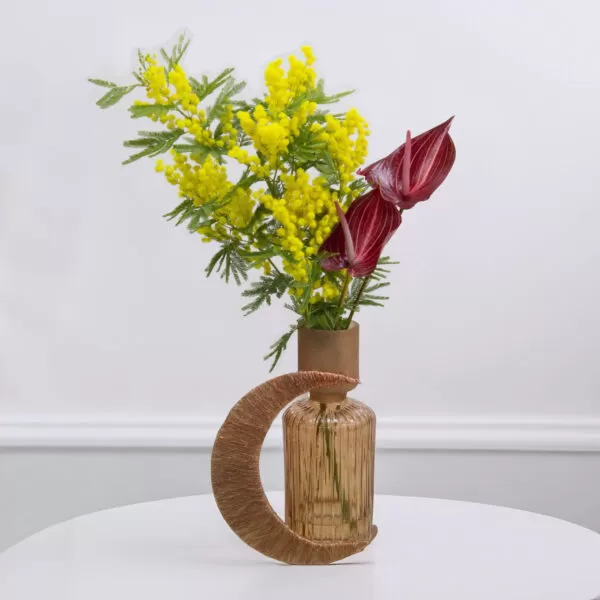 A tall, decorative glass vase with a narrow neck and a wider, rounded base, holding yellow mimosa flowers and red anthurium flowers, with a crescent design element made of textured material, placed on a white round table against a white wall.