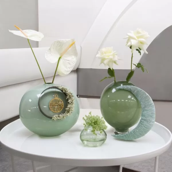 A light green, round vase with a ring and dried flowers, holding a white anthurium flower, a light green, round vase with a crescent design, holding two white roses, and a small, clear glass vase with green flowers, all on a white round table.