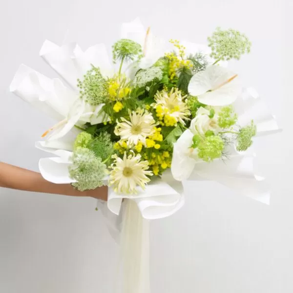 A close-up shot of a bouquet featuring white anthuriums, yellow chrysanthemums, green lady's mantle flowers, and yellow craspedia flowers, wrapped in white paper, held by a person's arm against a light background.