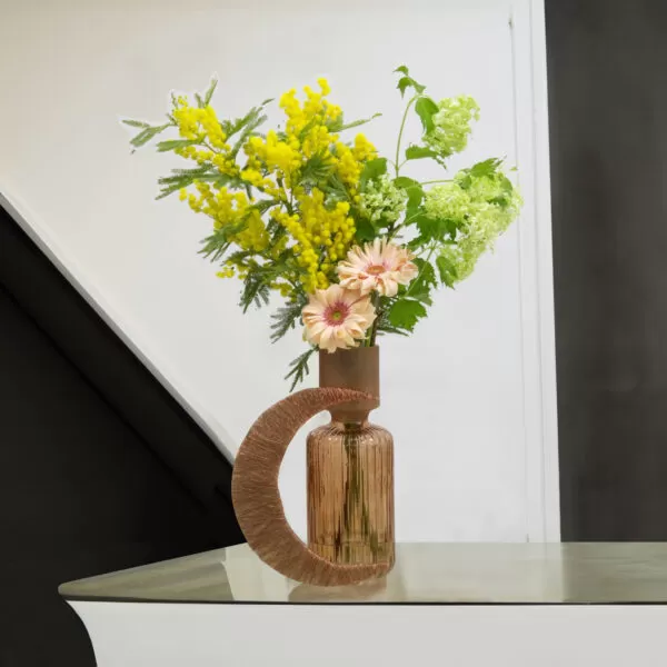 A tall, decorative glass vase with a narrow neck and a wider, rounded base, holding yellow mimosa flowers, pink gerberas, and green snowball flowers, with a crescent design element made of textured material, placed on a light-colored table against a white wall with a black diagonal stripe.