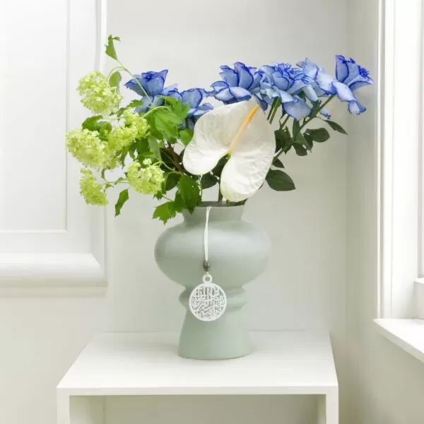 A light green, round vase with a tag hanging from it, holding blue roses, green snowball flowers, and a white anthurium flower, placed on a white shelf against a white wall with a window.