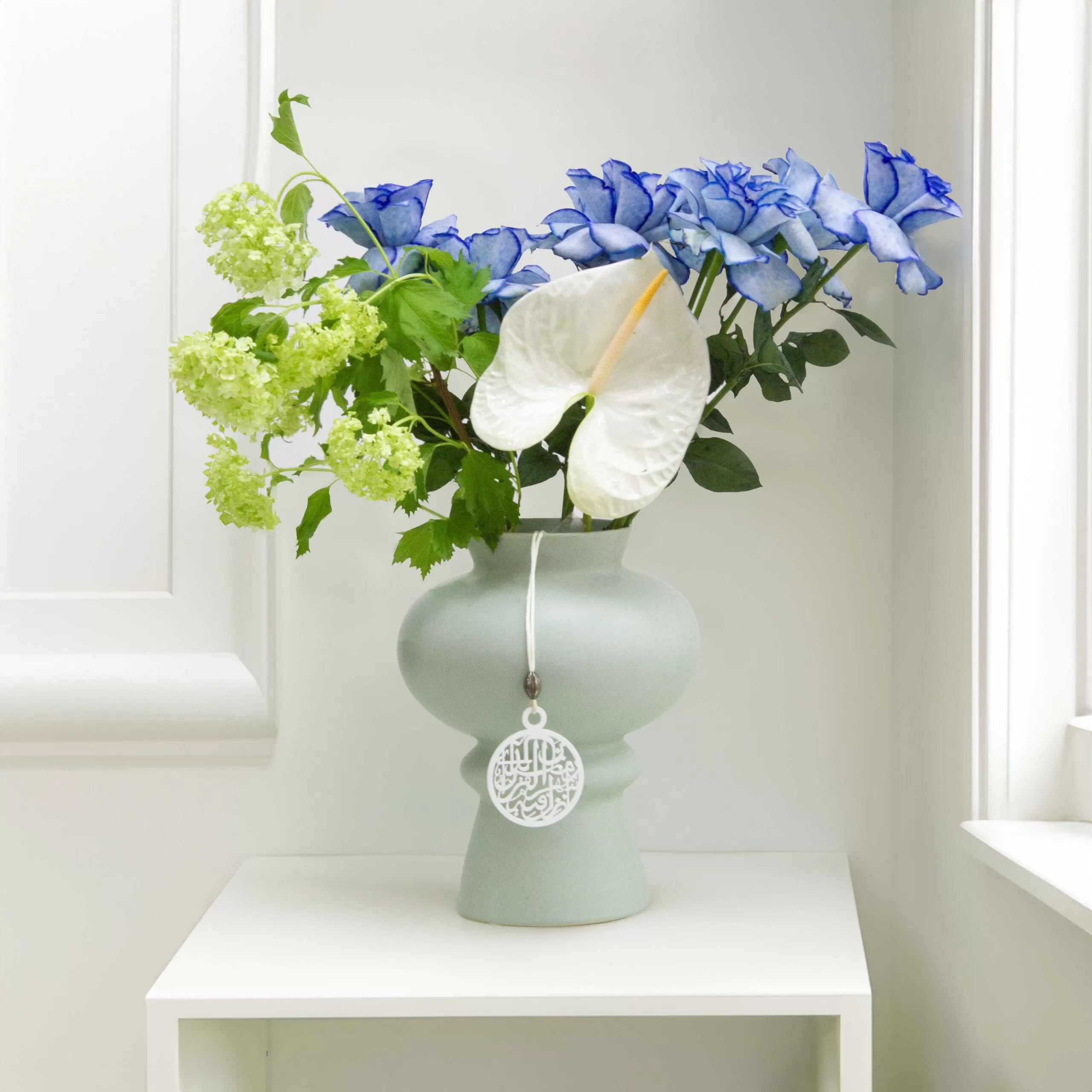 A light green, round vase with a tag hanging from it, holding blue roses, green snowball flowers, and a white anthurium flower, placed on a white shelf against a white wall with a window.