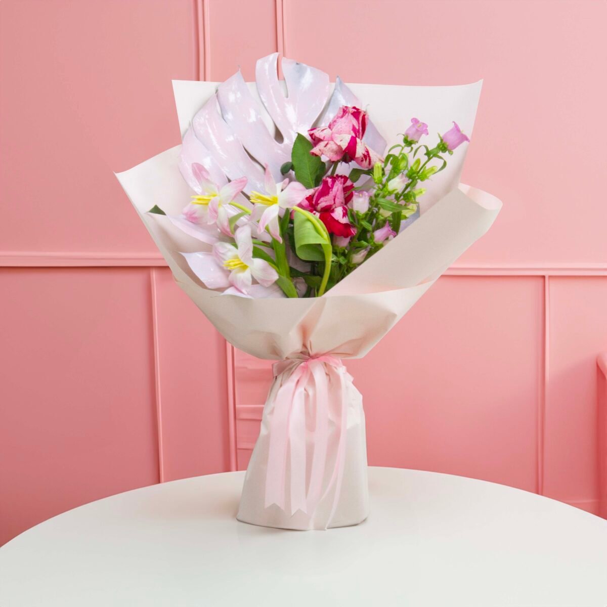 A close-up shot of a bouquet featuring pink and white tulips, small pink and purple flowers, and a large, white Monstera leaf, wrapped in light-colored paper and tied with a light pink ribbon, placed on a white round table against a pink panelled wall.