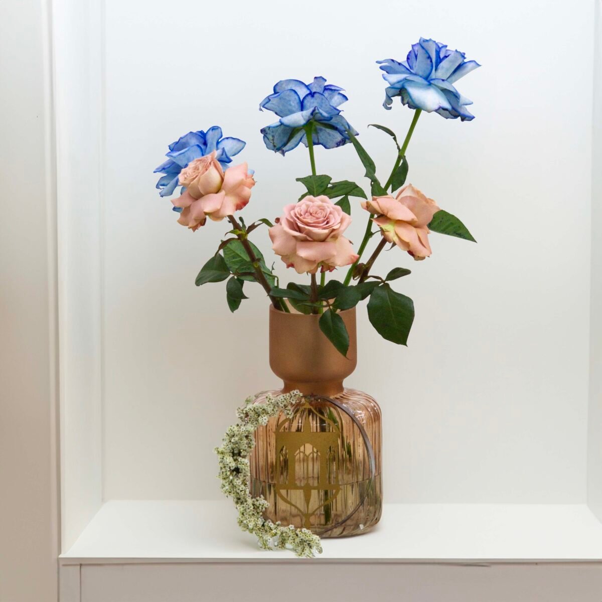 A decorative glass vase with a narrow neck and a wider, rounded base, holding three blue roses and three pink roses, with a floral ring of small white flowers around its base, placed on a white shelf against a white wall.