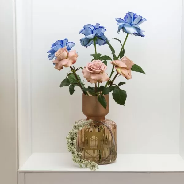 A decorative glass vase with a narrow neck and a wider, rounded base, holding three blue roses and three pink roses, with a floral ring of small white flowers around its base, placed on a white shelf against a white wall.
