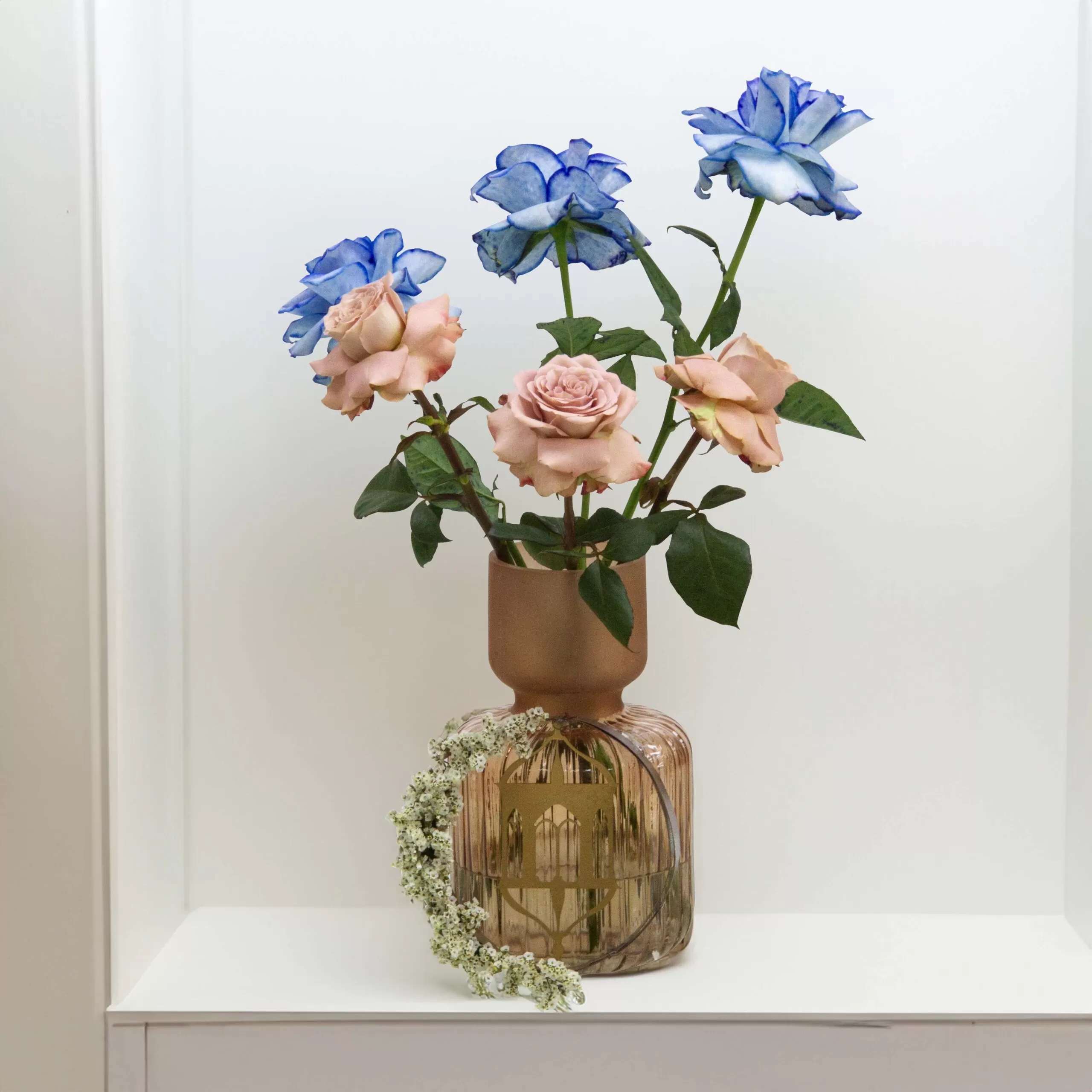 4 copy A decorative glass vase with a narrow neck and a wider, rounded base, holding three blue roses and three pink roses, with a floral ring of small white flowers around its base, placed on a white shelf against a white wall.