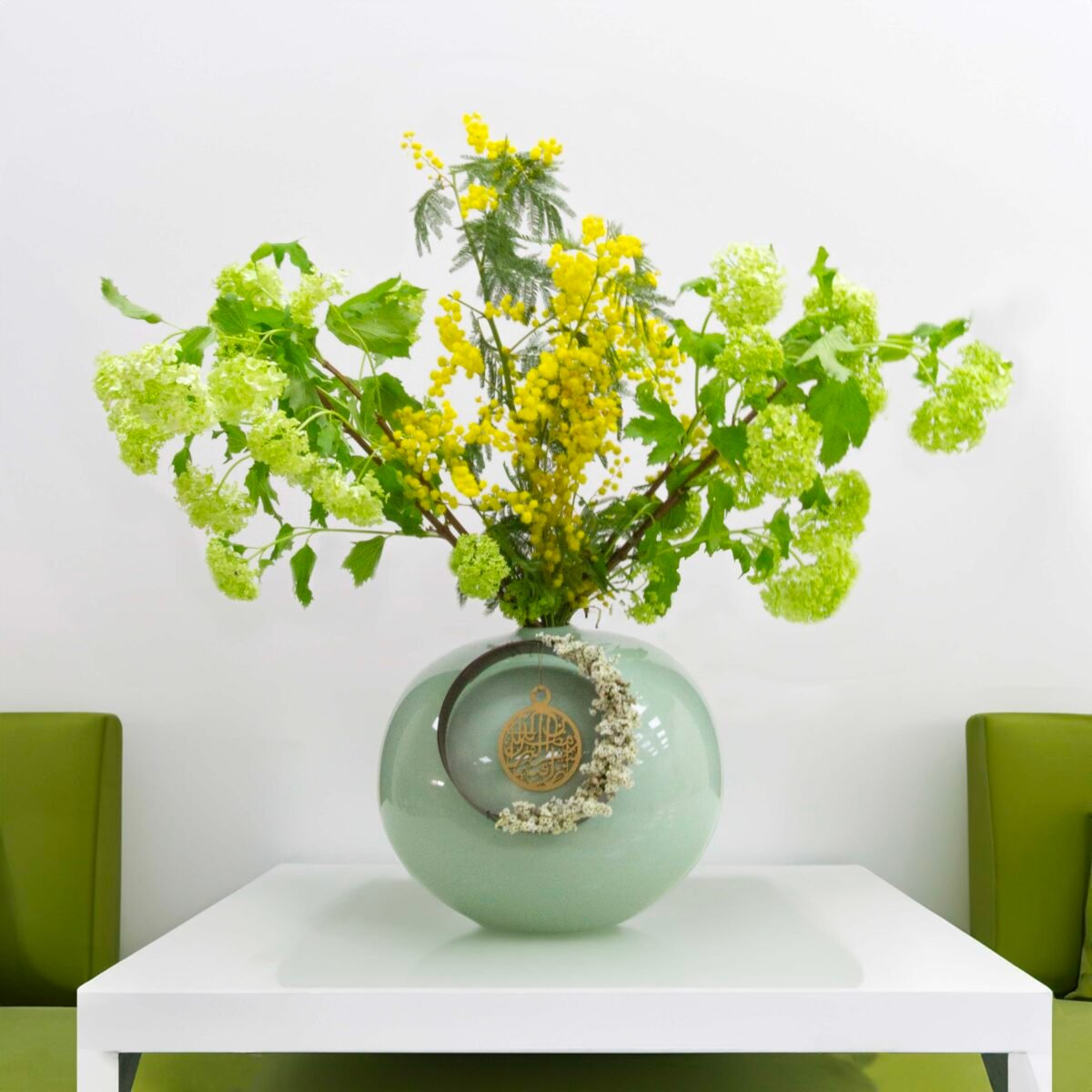 A light green, round vase with a ring and dried flowers, holding yellow mimosa flowers and green snowball flowers, placed on a white square table against a white wall with two green upholstered chairs.