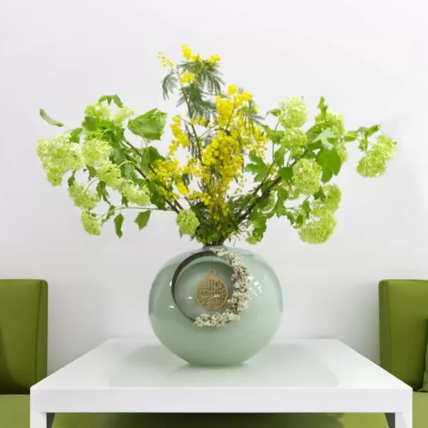 A light green, round vase with a ring and dried flowers, holding yellow mimosa flowers and green snowball flowers, placed on a white square table against a white wall with two green upholstered chairs.