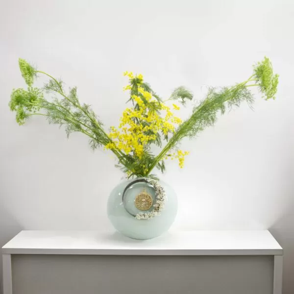 A light green, round vase with a ring and dried flowers, holding yellow mimosa flowers and green ammi flowers with feathery stems, placed on a white shelf against a white wall.