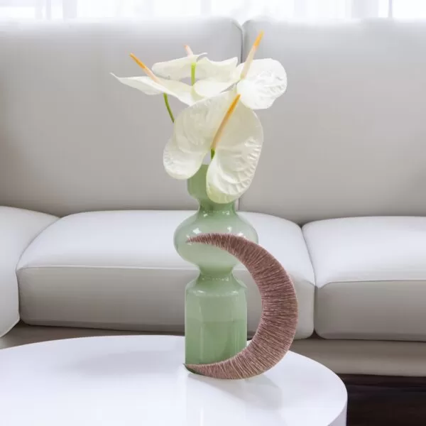 A light green, round vase with a crescent design made of textured material, holding white anthurium flowers with yellow stamens, placed on a white round table in front of a white, modern-style couch.