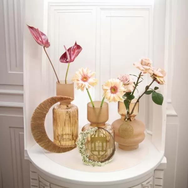 Three decorative glass vases with flowers: a tall, cylindrical vase with a crescent design holding a red anthurium, a shorter, cylindrical vase with a ring of flowers holding yellow gerberas, and a rounded vase holding pink roses, all arranged on a white round table with a decorative backdrop.