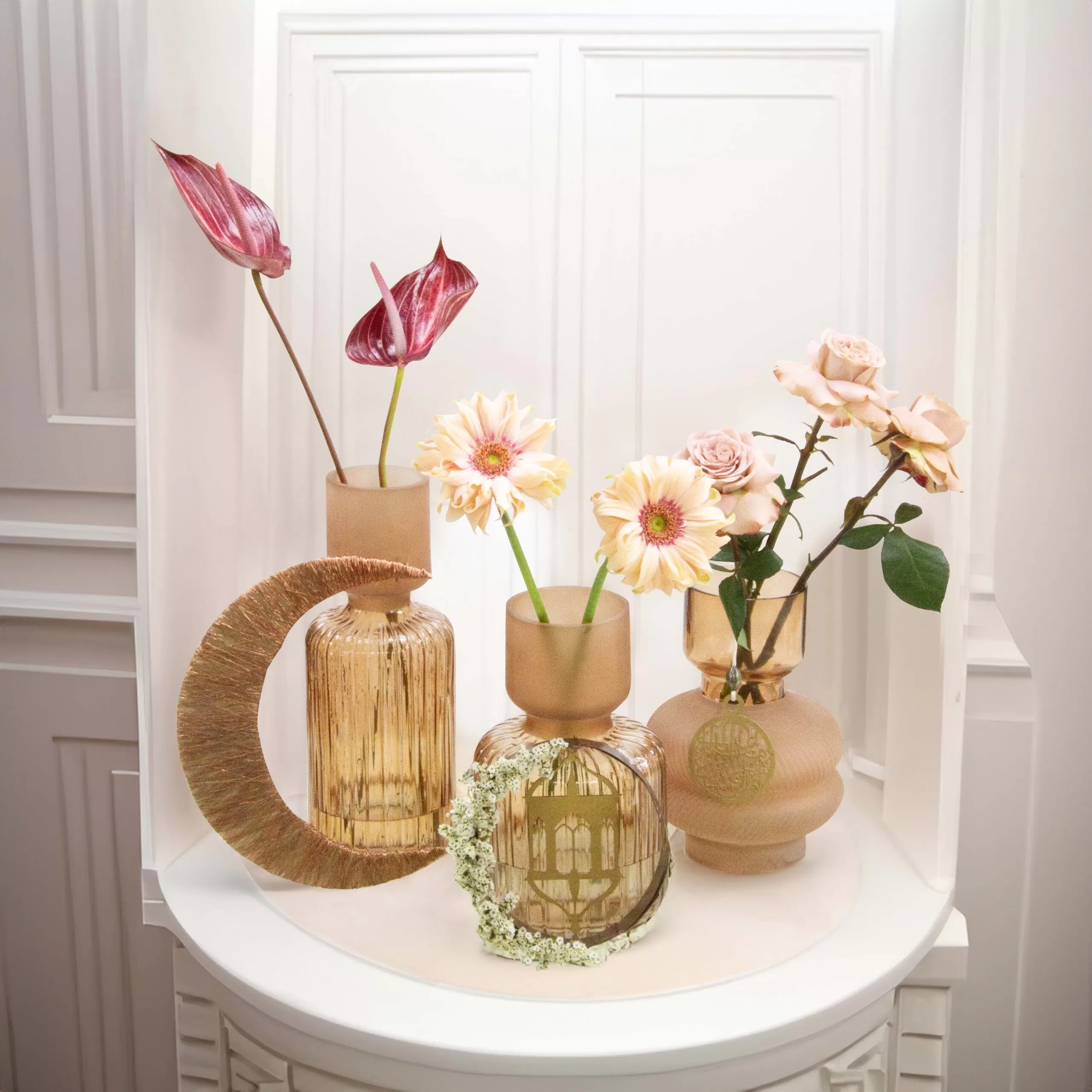 Three decorative glass vases with flowers: a tall, cylindrical vase with a crescent design holding a red anthurium, a shorter, cylindrical vase with a ring of flowers holding yellow gerberas, and a rounded vase holding pink roses, all arranged on a white round table with a decorative backdrop.