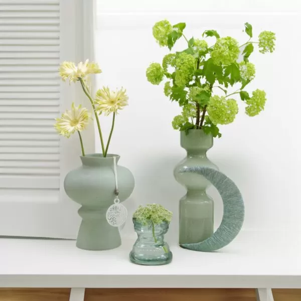 A light green, round vase with a ring and a tag, holding yellow flowers, a light green, round vase with a crescent design, holding green snowball flowers, and a small, clear glass vase with green flowers, all on a white shelf near a white window.