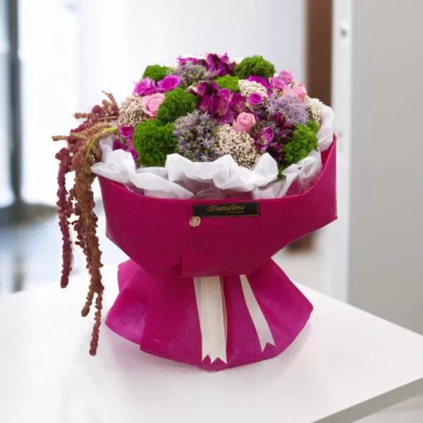 A bouquet of pink roses, purple allium, green dianthus, white astilbe, and burgundy amaranthus, wrapped in pink paper and tied with a white ribbon, placed on a white table with a blurred background. The bouquet also features a small black label with the word "Intraflora" on it.