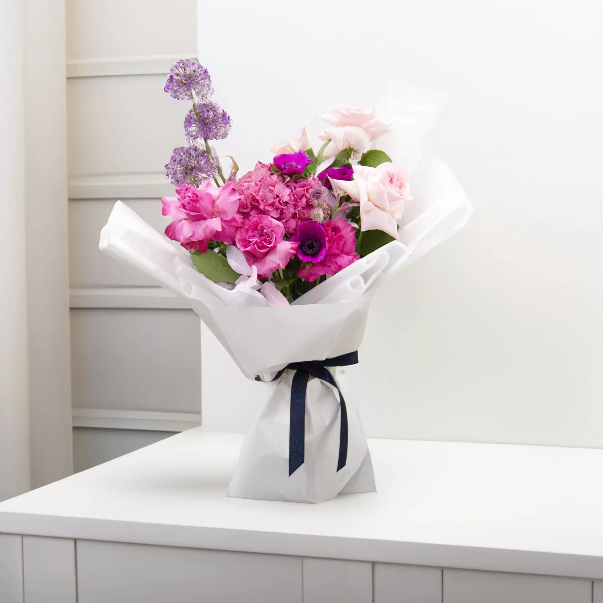 A bouquet featuring pink roses, purple allium flowers, and other pink and purple blooms, wrapped in white paper and tied with a dark blue ribbon, placed on a white table or counter.