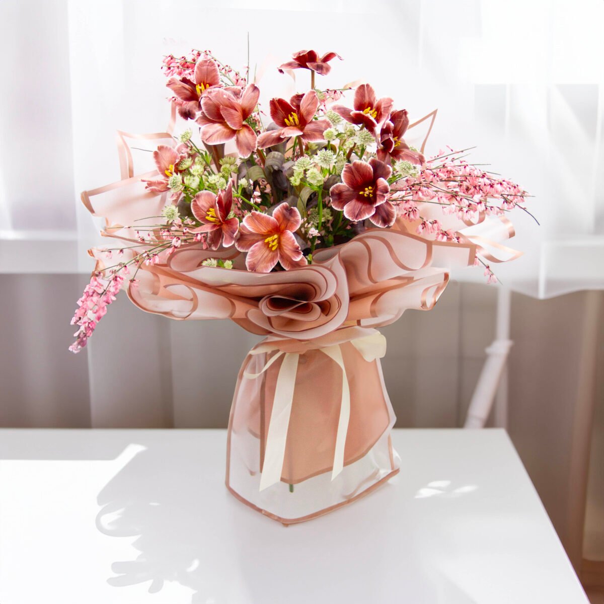 A bouquet of brown and pink lilies, pink cherry blossoms, and white baby's breath flowers, wrapped in pink fabric and tied with a pink ribbon, placed on a white table with a blurred background.