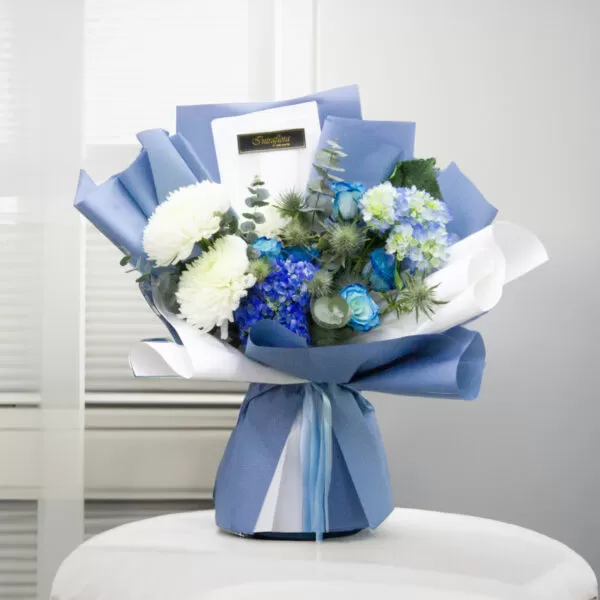 This image shows a bouquet of white chrysanthemums, blue roses, blue delphiniums, blue thistle, and green eucalyptus leaves, wrapped in blue and white paper and tied with a blue ribbon, placed on a white round table with a blurred background. The bouquet also features a white card with the word "Sunshine" on it.