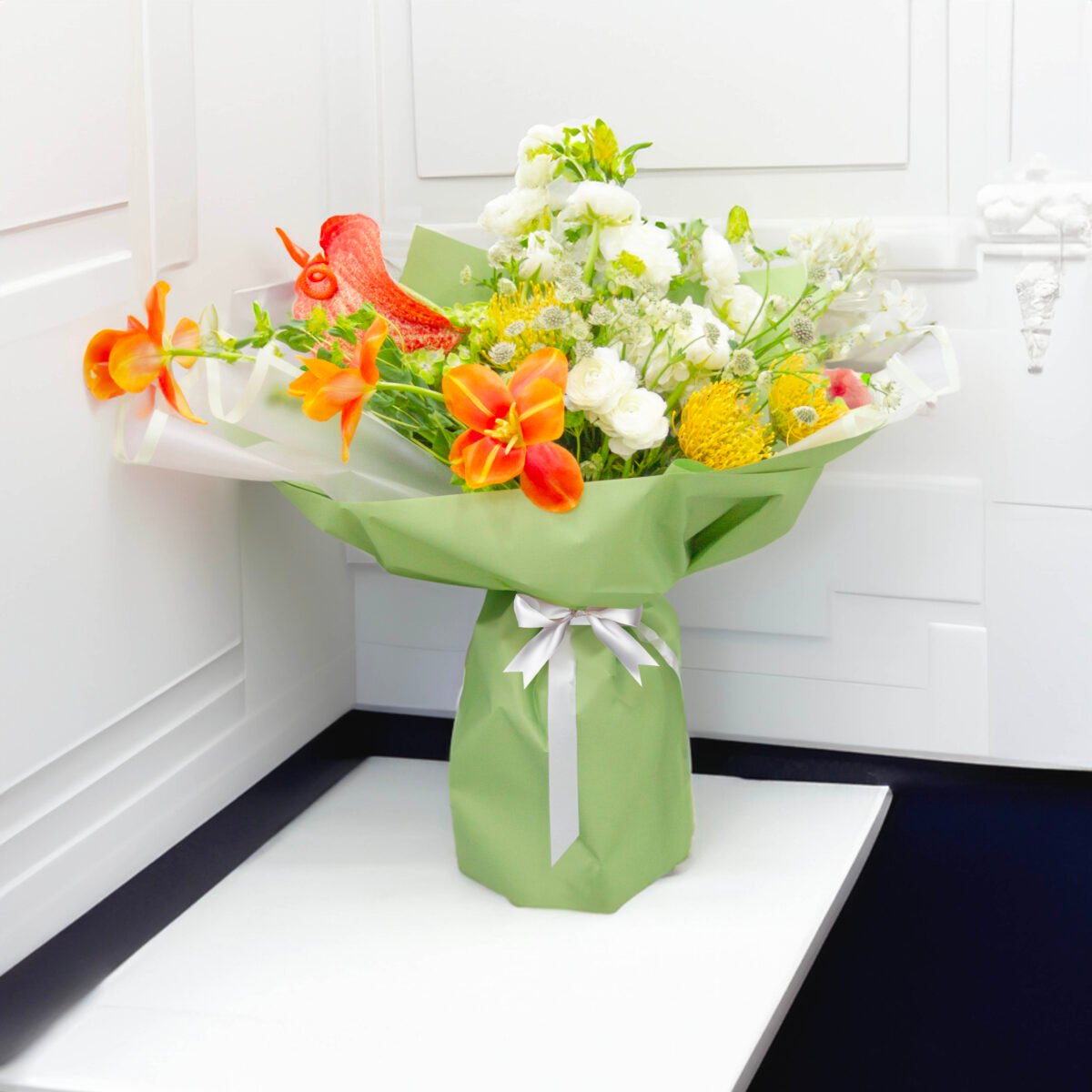 A bouquet of orange lilies, white roses, yellow craspedia, white delphinium, and white alstroemeria, wrapped in green paper and tied with a silver ribbon, placed on a white table with a white wall in the background.