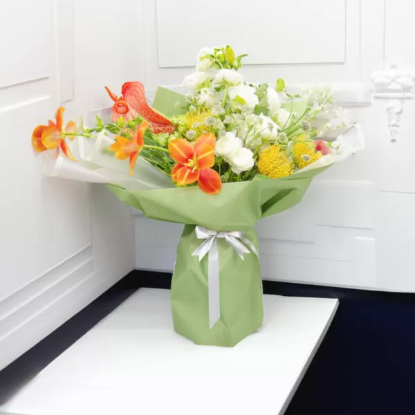 A bouquet of orange lilies, white roses, yellow craspedia, white delphinium, and white alstroemeria, wrapped in green paper and tied with a silver ribbon, placed on a white table with a white wall in the background.