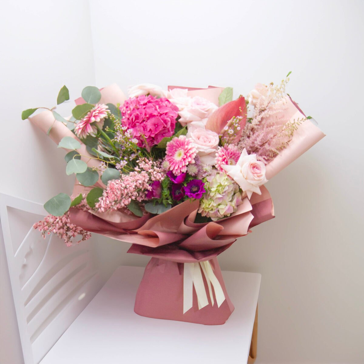 A bouquet featuring pink and white garden roses, pink hydrangeas, pink astilbe, and possibly peonies, wrapped in pink paper with a light pink ribbon, placed on a white table or counter.