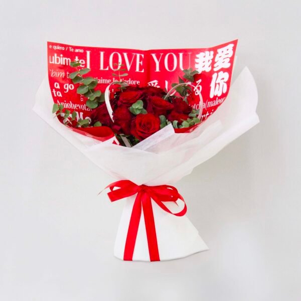 A bouquet of red roses wrapped in white paper with the phrase "I LOVE YOU" written in multiple languages, tied with a red ribbon, against a light background.