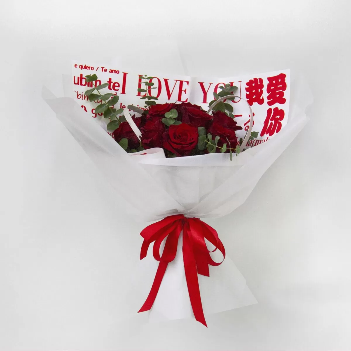 A bouquet of red roses wrapped in white paper with the phrase "I LOVE YOU" written in multiple languages, tied with a red ribbon, against a light background.