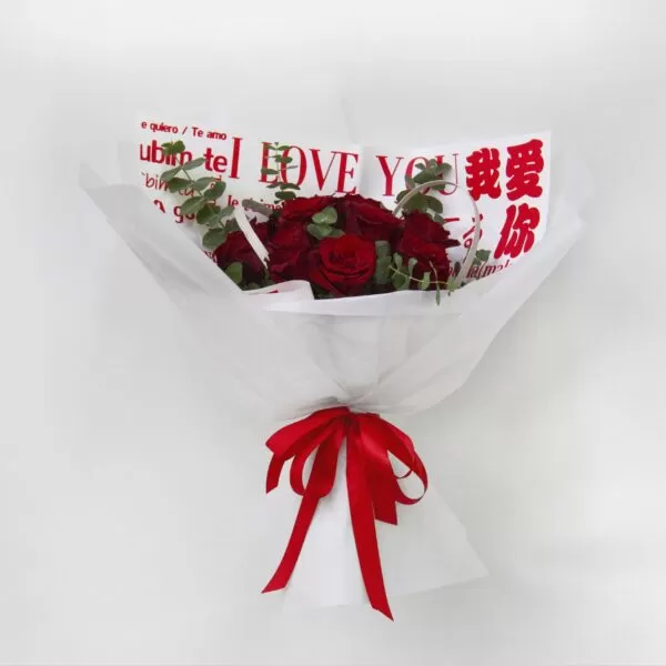 A bouquet of red roses wrapped in white paper with the phrase "I LOVE YOU" written in multiple languages, tied with a red ribbon, against a light background.